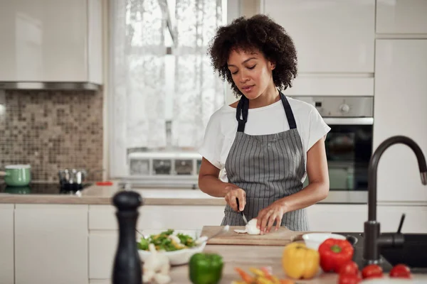Gorgeous blandad ras hemmafru i förkläde stående i köket och hugga svamp. På köksbänken finns alla typer av grönsaker. — Stockfoto