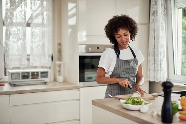 Gorgeous blandad ras hemmafru i förkläde stående i köket och hugga svamp. På köksbänken finns alla typer av grönsaker. — Stockfoto