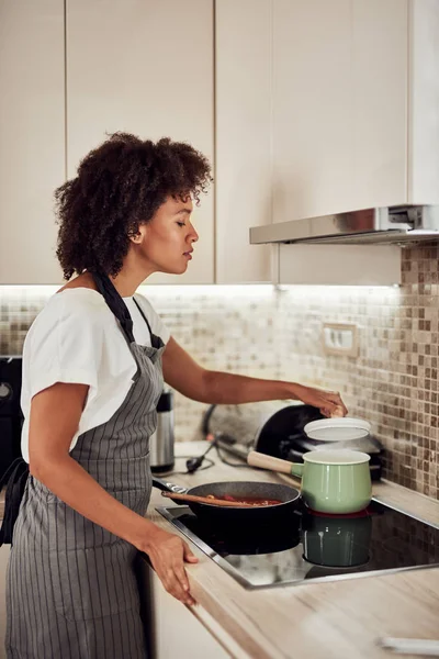 Mixed race woman in apron standing next to stove in domestic kitchen and making pasta and sauce.