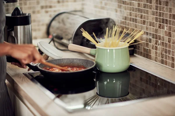 Смешанная расовая женщина в фартуке стоит рядом с плитой в домашней кухне и делает макароны и соус . — стоковое фото