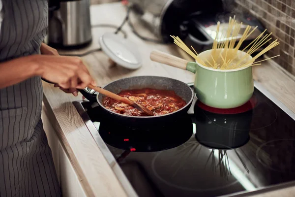 Расовая женщина в фартуке, стоящая рядом с плитой и перемешивающая томатный соус. На плите кастрюля и горшок со спагетти . — стоковое фото