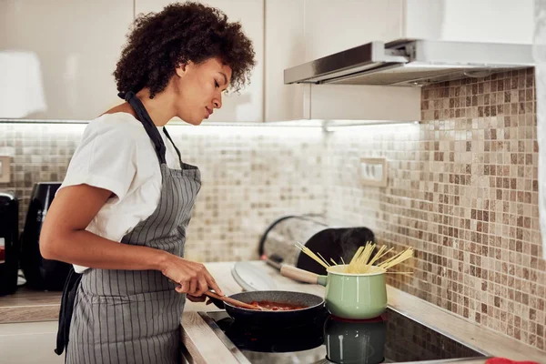 Mulher de raça mista em avental de pé ao lado do fogão e mexendo molho de tomate. No fogão estão panela e panela com espaguete . — Fotografia de Stock