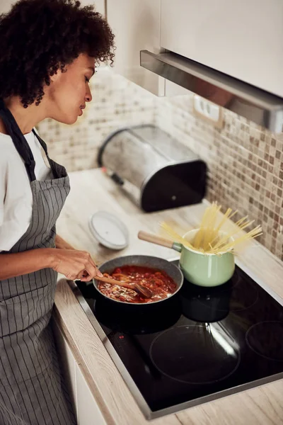 Mulher de raça mista em avental de pé ao lado do fogão e mexendo molho de tomate. No fogão estão panela e panela com espaguete . — Fotografia de Stock