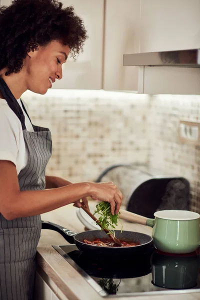 Mulher de raça mista em avental de pé ao lado do fogão, mexendo molho de tomate e adicionando foguete. No fogão é panela com massa fervente . — Fotografia de Stock