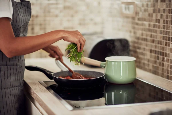 Mujer de raza mixta en delantal de pie junto a la estufa, revolviendo la salsa de tomate y añadiendo cohete. En la estufa es olla con pasta hirviendo . — Foto de Stock