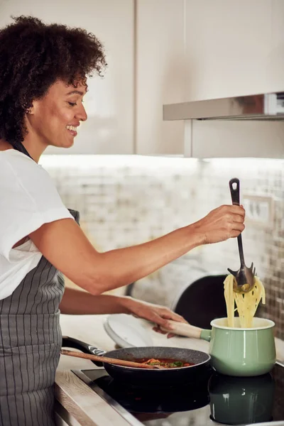Bela mulher de raça mista em avental de pé ao lado do fogão e fazendo espaguete . — Fotografia de Stock