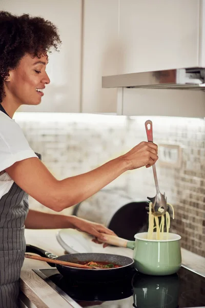 Bela mulher de raça mista em avental de pé ao lado do fogão e fazendo espaguete . — Fotografia de Stock