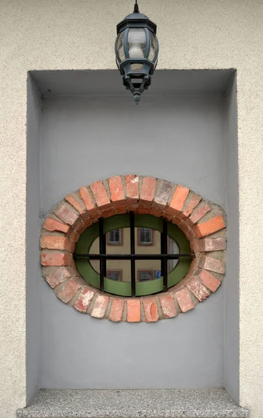 Oval window lined with brick and grate with reflection of house and lamp above it