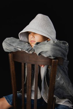 11 yaşında kapüşonlu süveter ve kot ceketli siyah bir arka planda tahta sandalyede oturan bir çocuk.