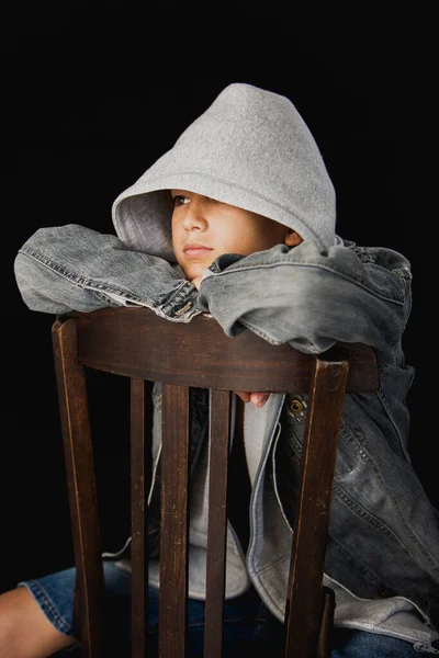 11 yaşında kapüşonlu süveter ve kot ceketli siyah bir arka planda tahta sandalyede oturan bir çocuk.