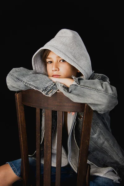 11 yaşında kapüşonlu süveter ve kot ceketli siyah bir arka planda tahta sandalyede oturan bir çocuk.
