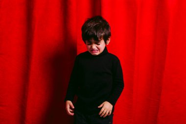 Üç yaşında bir çocuk, siyah giyinmiş, kırmızı perdeye karşı ağlıyor.