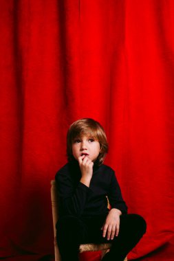 Beş yaşında, elleri ağzında, kırmızı bir perdeye yaslanmış düşünceli bir çocuk.