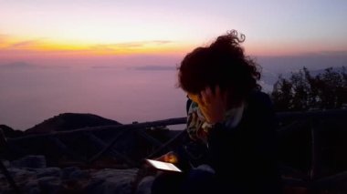 Pimonte, İtalya. Faito Dağı. 16 Ağustos 2020. Dağın tepesinde, önünde muhteşem bir günbatımı olan cep telefonunu kullanan genç bir kadın. Düşünceleri ve endişeleri olan bir kadın. Kafasını kaşıyan bir kadın.