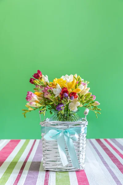 Bouquet of flowers in a glass vase placed inside a wicker wooden container on which a light blue ribbon is placed. The vase is placed on a table covered with a multicolored tablecloth against a green background.