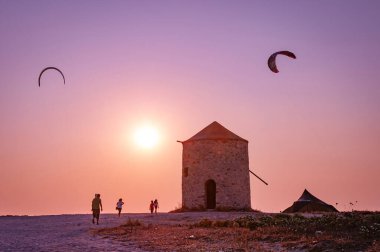 Lefkada, Yunanistan. 18 Ağustos 2011. İnsanlar gün batımında eski değirmenin yanındaki Milos plajına yürüyorlar. Gökyüzünde iki uçurtma sörfçüsünün uçurtmaları.