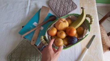 Masaya, kitapların ve diğer mutfak aletlerinin yanına yerleştirilmiş meyve sepetinden iki eriği attıktan sonra eriği seçen beyaz bir beyaz adamın eli..