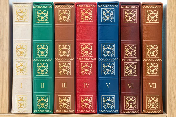 Leather-bound antique books, sorted from first to seventh volume on a wooden shelf with Roman numbers.