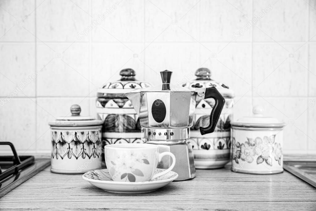 Italy. A cup of coffee and a coffee pot placed in front of pretty and colorful Vietri ceramic jars on the kitchen counter. Black and white image.