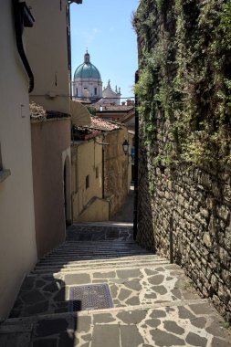 Brescia, Italy - 1ST AUGUST 2025 - Alley made of a staircase between a building and a stone wall with the dome of the Brescia basilica in the background on a sunny day