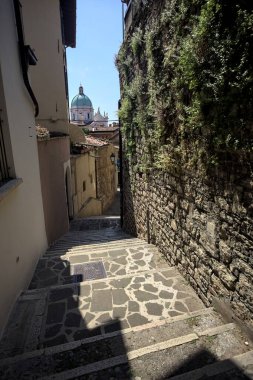 Brescia, Italy - 1ST AUGUST 2025 - Alley made of a staircase between a building and a stone wall with the dome of the Brescia basilica in the background on a sunny day