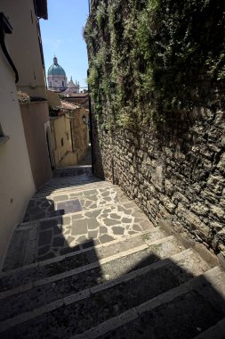 Brescia, Italy - 1ST AUGUST 2025 - Alley made of a staircase between a building and a stone wall with the dome of the Brescia basilica in the background on a sunny day