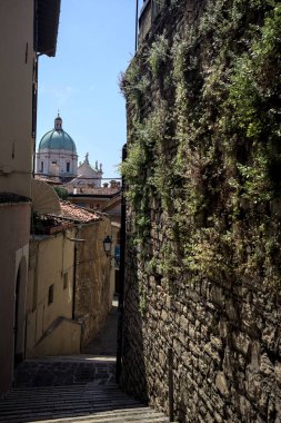 Brescia, Italy - 1ST AUGUST 2025 - Alley made of a staircase between a building and a stone wall with the dome of the Brescia basilica in the background on a sunny day