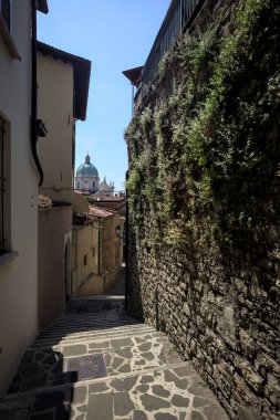 Brescia, Italy - 1ST AUGUST 2025 - Alley made of a staircase between a building and a stone wall with the dome of the Brescia basilica in the background on a sunny day