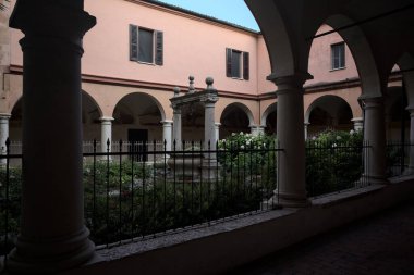 Cloister bordered by bushes with a well in the middle of it seen from the porch at sunset