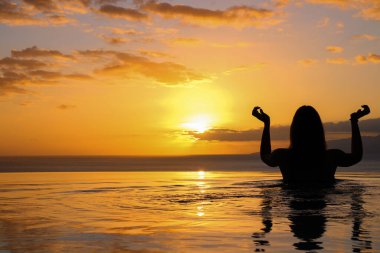 Kız sırtı havuza dönük duruyor, elleri bir Hint dansı gibi katlanmış, okyanusta batan güneşin arka planına karşı. Gia de Isora. Tenerife. İspanya