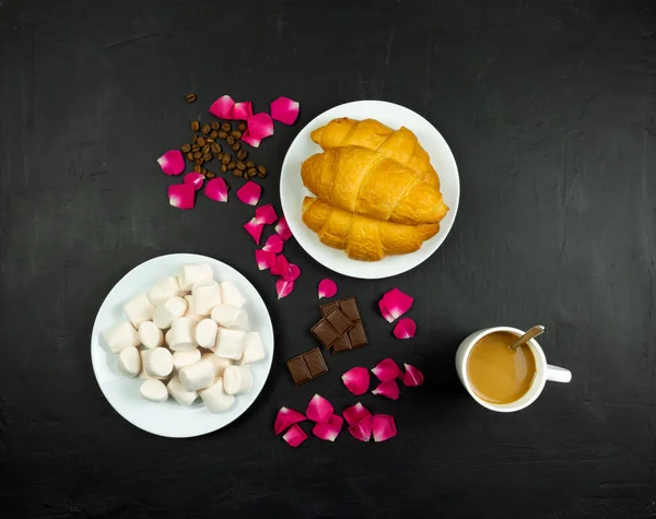 Appetizing croissants, marshmallows, chocolate and rose petals on a dark background. Top view