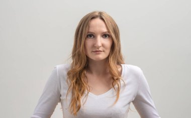 Beyaz tişört giyen, gri arka planda, sakince ve dikkatlice kameraya bakan, beyaz saçlı, ciddi odaklanmış beyaz bir kadının stüdyo portresi.