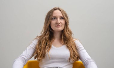 Sakin, ciddi, odaklanmış, beyaz tişört giyen, sakince ve dikkatle yana bakan, gri arka plan üzerinde kolları olan, uzun altın sarısı saçlı beyaz kadın.