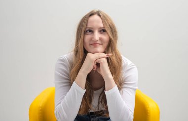 Düşünceli genç beyaz bir kadının portresi. Sarı koltukta oturmuş, kolları çenesinde, stüdyo çekimlerinde ciddi bir şekilde yana bakıyor.