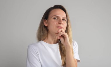 Kafası karışmış, güvenilmez yüzlü, gri stüdyo duvarında izole edilmiş, kuşkulu, kuşkulu, kameraya bakan, şüpheli, sarışın, beyaz kadın.