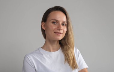 Kendine güvenen, gülümseyen genç Avrupalı bir kadın, beyaz tişörtlü, kameraya sakin ve cana yakın bakan, gri stüdyo arka planında izole edilmiş başarılı bir kadın.