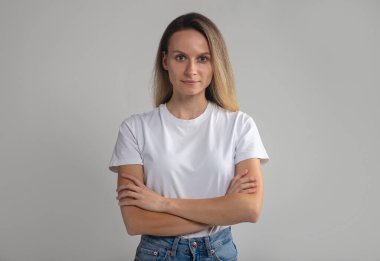 Kendine güveni tam, gülümseyen, kollarını kavuşturmuş, beyaz tişörtlü, sakin bir şekilde kameraya bakan, gri stüdyo arka planında izole edilmiş başarılı bir kadın.