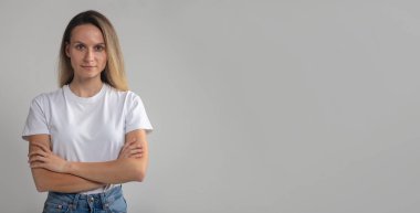 Kendine güveni tam, gülümseyen güzel Avrupalı kadın kollarını kavuşturmuş, beyaz tişörtlü, sakince kameraya bakıyor, başarılı bir kadın stüdyoda poz veriyor.