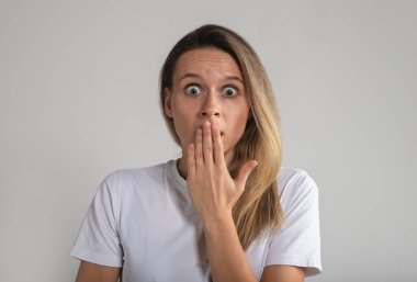 Ağzını avuç içiyle kapatan neşeli genç bayana ne mutlu bir sürpriz. Gri arka planda izole edilmiş bir kadın, stüdyoda sarışın Avrupalı bir kadın.