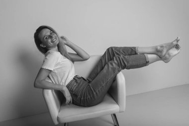 Çekici, mutlu, genç bir kadın. Üzerinde bacakları olan bir sandalyede rahatlıyor. Gülümsüyor ve kameraya bakıyor.