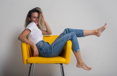 Güzel genç bir kadın sarı sandalyede oturuyor ya da uzanıyor. Bacakları havada ve gururla kameraya bakıyor. Stüdyo çekimi.