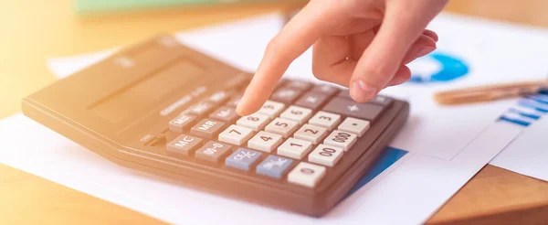close up female accountant or banker hand making calculations sitting at office table with documents with tables on desk. Savings, finances and economy concept, counting on calculator, banner