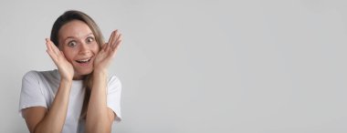 Genç, komik, güzel bir kadın hoş bir şekilde ellerini yerleştirir ve gri arka planda şaşırmış gibi yapar. kopyalama alanı, afiş