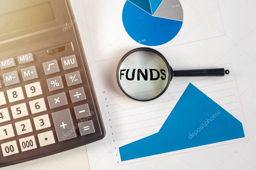 FUNDS Word through magnifying glass on white table with calculator and blue charts. Income, expenses, tax, financial data.