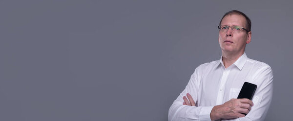 Студия снимает грустного старшего бизнесмена, думающего, глядя прямо и держа мобильный телефон в руке на сером фоне. копировальное пространство, баннер