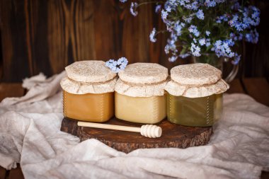 Üç kavanoz taze bal farklı renklerde beyaz kahverengi ve sarı bir tahta testerenin üzerinde kahverengi bir masada kahverengi bir fonda mavi çiçeklerin yanında kahverengi bir masa örtüsü.