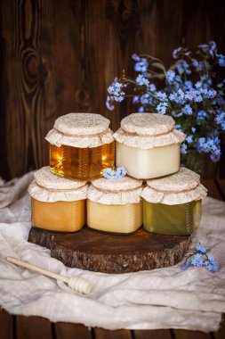 Çiğ organik ballı kavanoz piramidi. Bal sarısı, kahverengi, birbirinin üstünde duruyor. Tahta bir testere üzerinde keten bir masa örtüsü üzerinde kahverengi ahşap bir masada mavi çiçeklerin yanında.