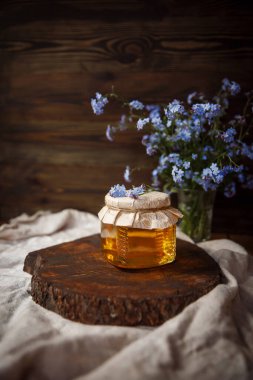 Kahverengi ahşap bir kesikte taze sunny ham bal kavanozu. Kapağının üstünde çiçekler vardı. Kapağı iple sarılmış. Kahverengi bir sehpanın üzerindeki keten kumaş üzerinde kahverengi bir ahşap arka planda mavi çiçeklerin yanında. Güzel, şık, lezzetli, tatlı, lezzetli kırsal... 