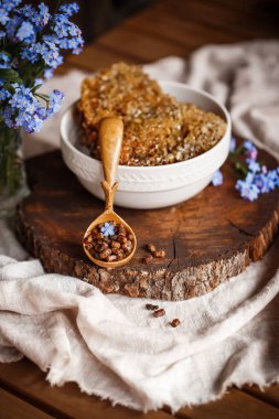 Ahşap kahverengi kaşık arı ürünleri ile Perga arı parşömeninin yanında bal ve ahşap bir kahverengi kesikle keten bir masa örtüsü üzerinde mavi çiçekler ve parşömenin üzerinde bir çiçek.