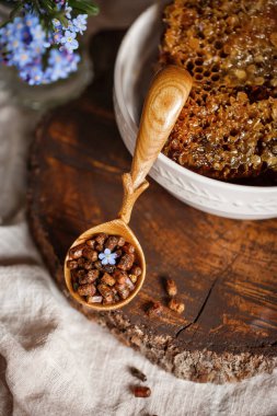 Ahşap kahverengi kaşık arı ürünleri ile Perga arı parşömeninin yanında bal ve ahşap bir kahverengi kesikle keten bir masa örtüsü üzerinde mavi çiçekler ve parşömenin üzerinde bir çiçek.